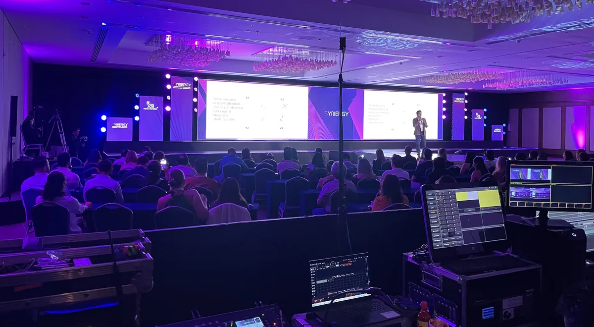 Salón con público sentado, escenario iluminado y consola en primer plano